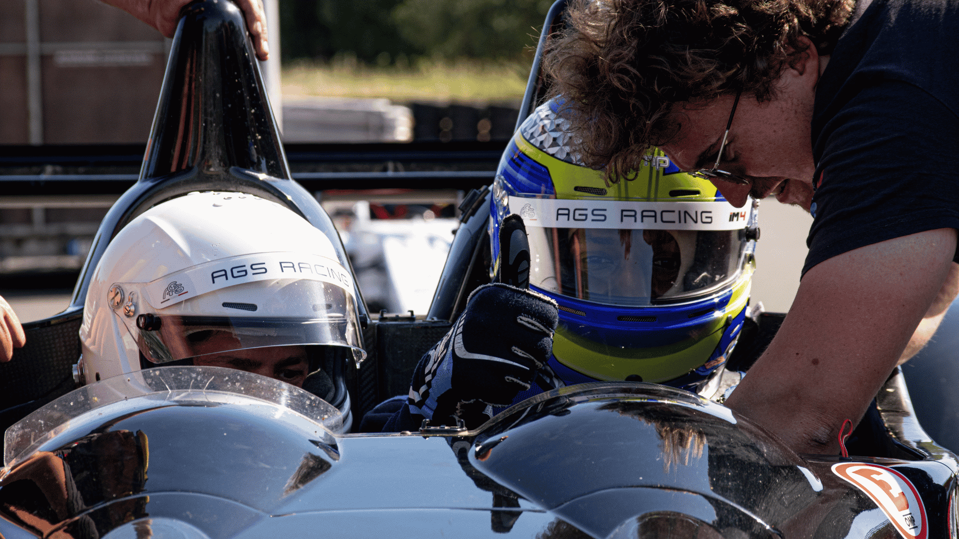 Twee coureurs met AGS Racing-helmen in de cockpit van een tweezitter Formule 1, tijdens briefing door de instructeur